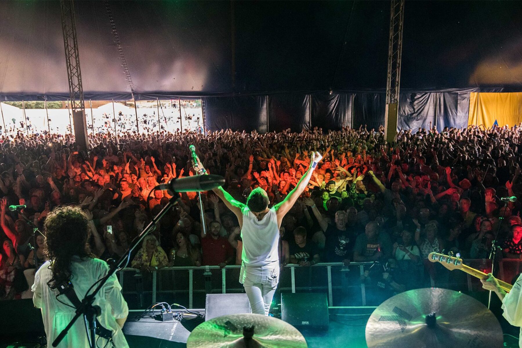 View from behind the drum kit looking out at a packed festival tent crowd with arms raised